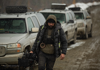 Photo: Adam Al-Samarea in a muddy field where cars are lined up alongside him. He is holding a camera in his right hand, wearing a hoodie and water proof jacket, it looks cold and wet.