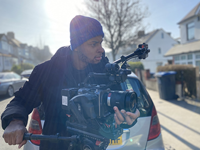 Photo: Dominic Christopher, a young black man with a short beard, is stood behind a video camera on a tripod in the middle of a residential street on a sunny winter's day.