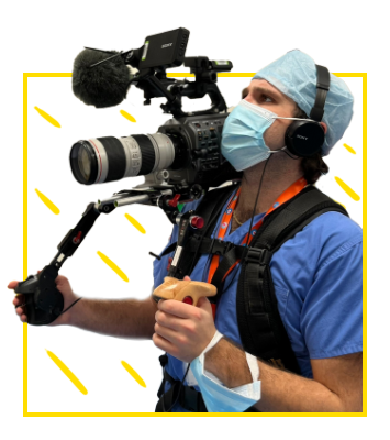 Headshot: Adam Al-Samarea – photo of a man in medical scrubs and wearing a face mask, holding a large broadcast camera on a rig over his right shoulder. 