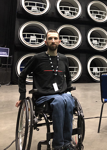 Photo: Stefan, a young white man with dark hair and beard, is sitting in a wheelchair on the set of a quiz show pilot. He is positioned in front of a large structure which will seat the contestants.